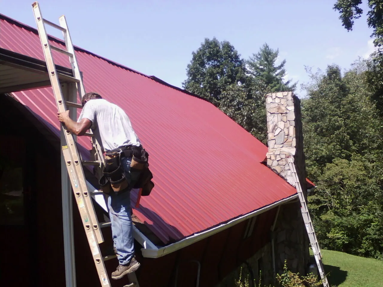 Roofer on a ladder beside a red metal roof and stone chimney — Soffit & Fascia Repair by Roofing Brothers in Lewisberry